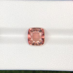 May include: A cushion-cut gemstone with a peach-pink hue. The gem is set against a white background, showcasing its facets and internal structure. The stone's color ranges from light to medium peach, with subtle variations.