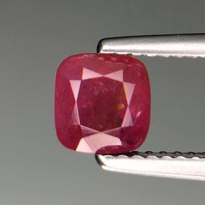 Puede incluir: Una gema de rubí con corte de cojín, sujeta con pinzas. El rubí es de color rojo intenso con facetas internas que crean un patrón de estrella. La gema se encuentra sobre un fondo gris neutro.