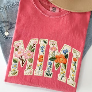 May include: A coral-colored t-shirt with the embroidered word "MIMI" in a floral design. The letters are outlined in a cream color and filled with pink, orange, and blue flowers. The shirt is laid on top of blue jeans and a tan hat.