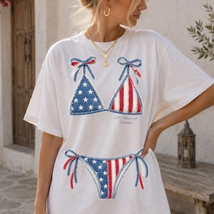 May include: White t-shirt featuring a patriotic bikini design in red, white, and blue. The bikini top has a star-spangled triangle and striped design. The matching bottom is also visible. The text "250 Years of Freedom" is printed below the bikini top.