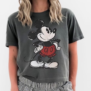May include: Dark gray t-shirt featuring a distressed graphic of Mickey Mouse in his classic pose. Mickey is wearing red shorts, white gloves, and shoes. The shirt is paired with black and white checkered shorts.