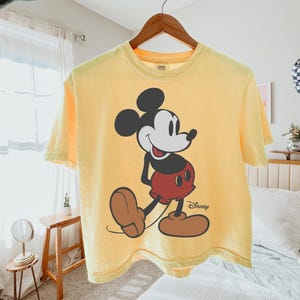 May include: Yellow t-shirt featuring a large graphic of Mickey Mouse. The cartoon character is depicted in black, white, red, and brown. The Disney logo is printed in black on the lower right side of the shirt.