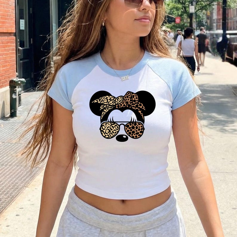 Op de afbeelding: Wit en lichtblauw crop top met korte mouwen met een afbeelding van een stripfiguur met een luipaardprint strik, zonnebril en een zwarte neus. De top heeft een baseball-shirtstijl met lichtblauwe mouwen.