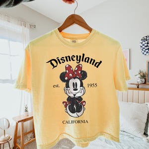 K&ouml;nnte beinhalten: Hellgelbes T-Shirt mit einem Minnie Mouse-Motiv mit roter Schleife und dem Schriftzug "Disneyland" &uuml;ber der Figur. Das Shirt enth&auml;lt auch den Text "est. 1955" und "CALIFORNIA" darunter.
