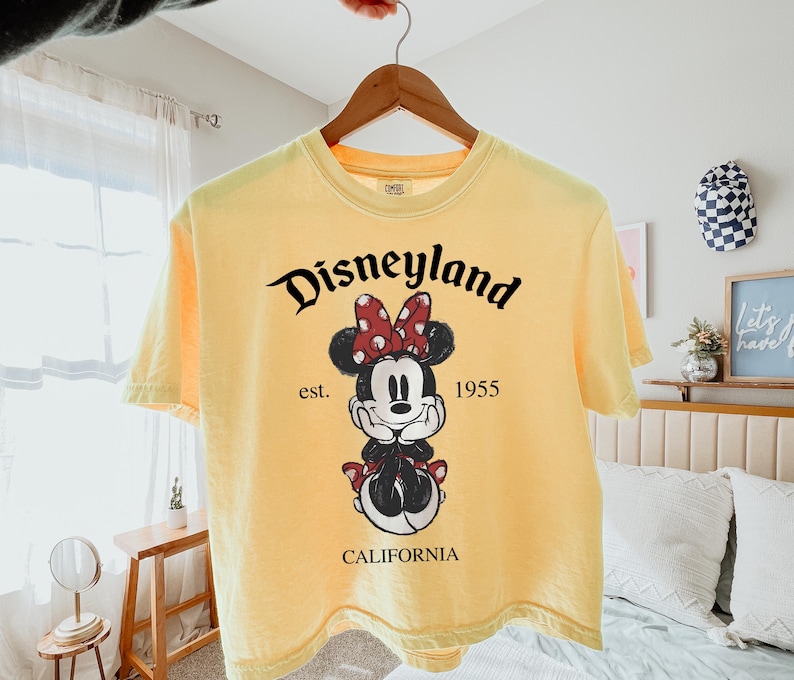 K&ouml;nnte beinhalten: Hellgelbes T-Shirt mit einem Minnie Mouse-Motiv mit roter Schleife und dem Schriftzug "Disneyland" &uuml;ber der Figur. Das Shirt enth&auml;lt auch den Text "est. 1955" und "CALIFORNIA" darunter.