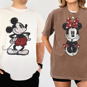 以下が含まれることがあります： ディズニーキャラクターが描かれたTシャツ2枚。1枚はクリーム色で、ミッキーマウスのグラフィックが黒、赤、白で描かれています。もう1枚は茶色で、ミニーマウスのグラフィックが黒、赤、白で描かれています。どちらのシャツも半袖です。