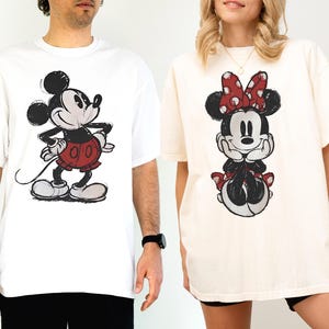 以下が含まれることがあります： 白とクリーム色の2枚のオーバーサイズTシャツ。それぞれにミッキーとミニーマウスのスケッチ風漫画が描かれています。ミッキーは赤いショートパンツ、ミニーは赤い水玉模様のリボンを着用。どちらのシャツもゆったりとしたフィット感です。
