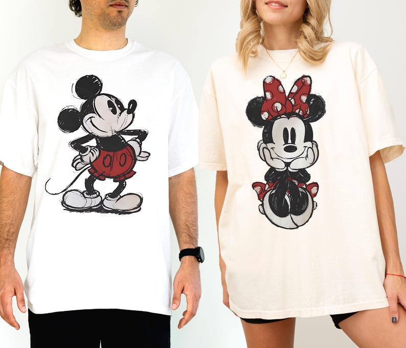 以下が含まれることがあります： 白とクリーム色の2枚のオーバーサイズTシャツ。それぞれにミッキーとミニーマウスのスケッチ風漫画が描かれています。ミッキーは赤いショートパンツ、ミニーは赤い水玉模様のリボンを着用。どちらのシャツもゆったりとしたフィット感です。
