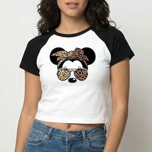 Op de afbeelding: Witte crop top met zwarte raglanmouwen met een afbeelding van een muis met een luipaardprint strik, zonnebril en oren. De muisafbeelding heeft een zwarte neus en draagt een zonnebril met luipaardprint.