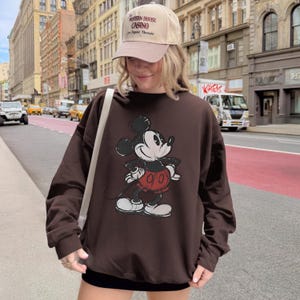 May include: A brown sweatshirt featuring a distressed Mickey Mouse graphic in black, white, and red. The sweatshirt is paired with a beige baseball cap with text. The image is taken outdoors with a city street in the background.