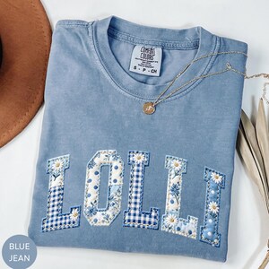 May include: A blue jean colored t-shirt with the word "LOLLI" appliqued across the front. The letters are a mix of floral, polka dot, and gingham patterns. A gold necklace with a pendant is draped across the shirt.