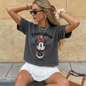 K&ouml;nnte beinhalten: Dunkelgraues T-Shirt mit einem Disneyland-Motiv mit Minnie Mouse. Das Shirt tr&auml;gt den Text "Disneyland est. 1955 CALIFORNIA". Das Shirt wird mit einem wei&szlig;en Rock kombiniert.