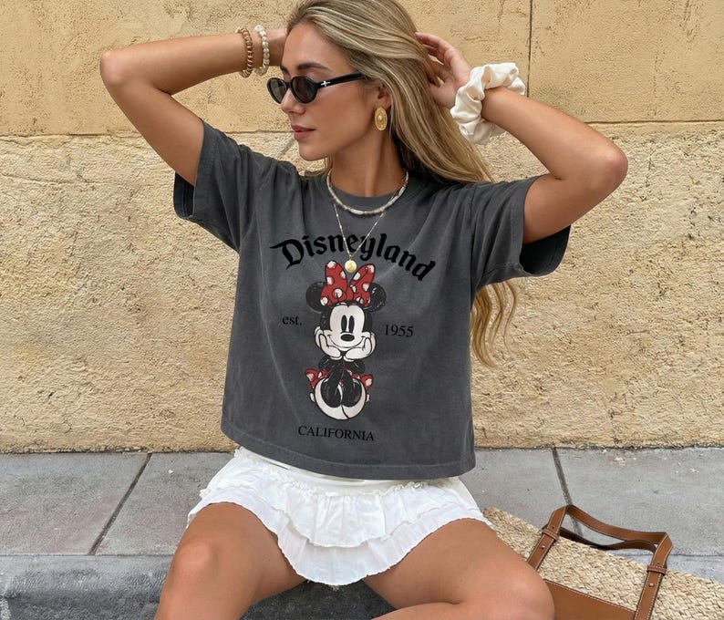 K&ouml;nnte beinhalten: Dunkelgraues T-Shirt mit einem Disneyland-Motiv mit Minnie Mouse. Das Shirt tr&auml;gt den Text "Disneyland est. 1955 CALIFORNIA". Das Shirt wird mit einem wei&szlig;en Rock kombiniert.