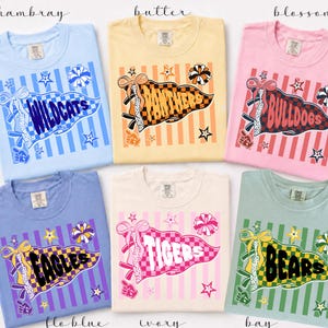 May include: Six short-sleeve crew neck t-shirts in light blue, yellow, pink, periwinkle, ivory, and sage green. Each shirt features a graphic of a pennant flag with a team name, including Wildcats, Panthers, Bulldogs, Eagles, Tigers, and Bears.