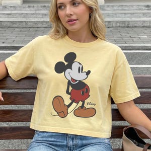 May include: Yellow t-shirt featuring a large graphic of Mickey Mouse in a classic pose. The cartoon character is depicted in black, red, and brown. The Disney logo is printed below the character. The shirt has short sleeves and a relaxed fit.