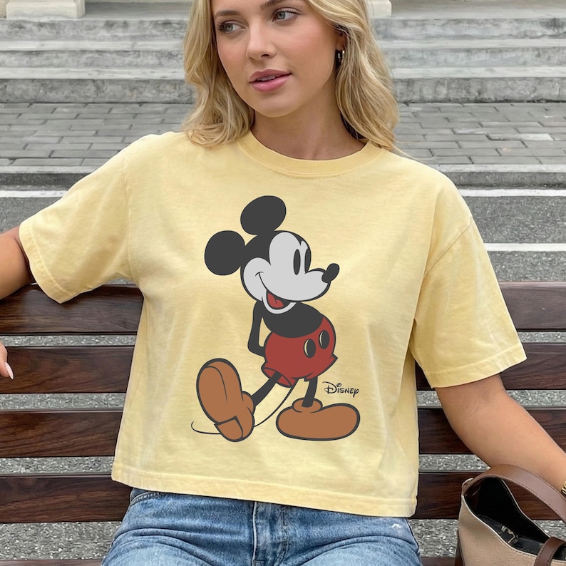 May include: Yellow t-shirt featuring a large graphic of Mickey Mouse in a classic pose. The cartoon character is depicted in black, red, and brown. The Disney logo is printed below the character. The shirt has short sleeves and a relaxed fit.