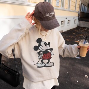 May include: A beige sweatshirt featuring a classic Mickey Mouse graphic in black, red, and white. The person is wearing a brown baseball cap with "La Paris" text and holding a black purse and a coffee drink.
