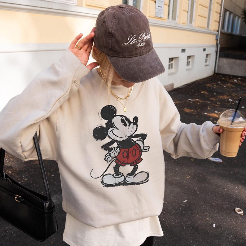 May include: A beige sweatshirt featuring a classic Mickey Mouse graphic in black, red, and white. The person is wearing a brown baseball cap with "La Paris" text and holding a black purse and a coffee drink.