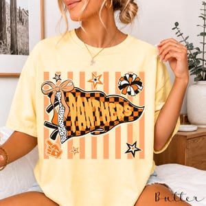 May include: A pale yellow t-shirt featuring a graphic print of a pennant flag with a black and orange checkerboard pattern. The word PANTHERS is written in orange letters on the flag, accompanied by a bow, a pom-pom, stars, and a foam finger.