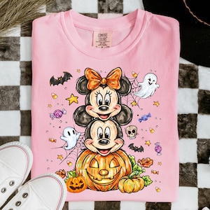 May include: A light pink t-shirt featuring a graphic print of Mickey and Minnie Mouse stacked on top of a large jack-o'-lantern. The design includes small ghosts, bats, a skull, candy, stars, and spiderwebs in orange, purple, and white colours.