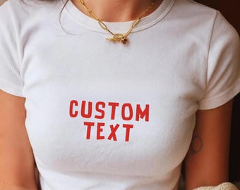 May include: White t-shirt with the red text "CUSTOM TEXT". The shirt has a crew neck and short sleeves. A gold-colored chain necklace with a toggle clasp is worn around the neck.