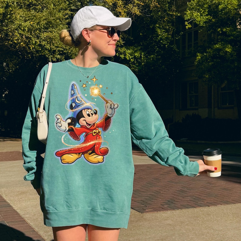 May include: Teal-colored sweatshirt featuring a graphic of Mickey Mouse dressed as a wizard, holding a glowing wand. The design includes a blue wizard hat with stars and a red robe. The sweatshirt has long sleeves and a relaxed fit.
