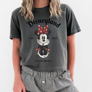 K&ouml;nnte beinhalten: Dunkelgraues T-Shirt mit Minnie Mouse-Grafik, roter Schleife und dem Text "Disneyland California est. 1955". Das T-Shirt hat eine lockere Passform.
