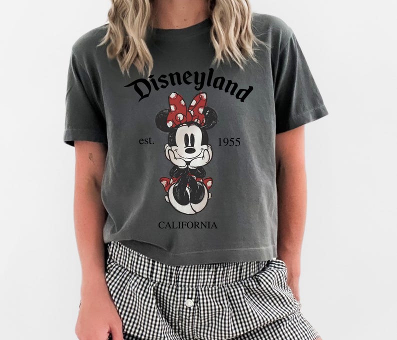 K&ouml;nnte beinhalten: Dunkelgraues T-Shirt mit Minnie Mouse-Grafik, roter Schleife und dem Text "Disneyland California est. 1955". Das T-Shirt hat eine lockere Passform.