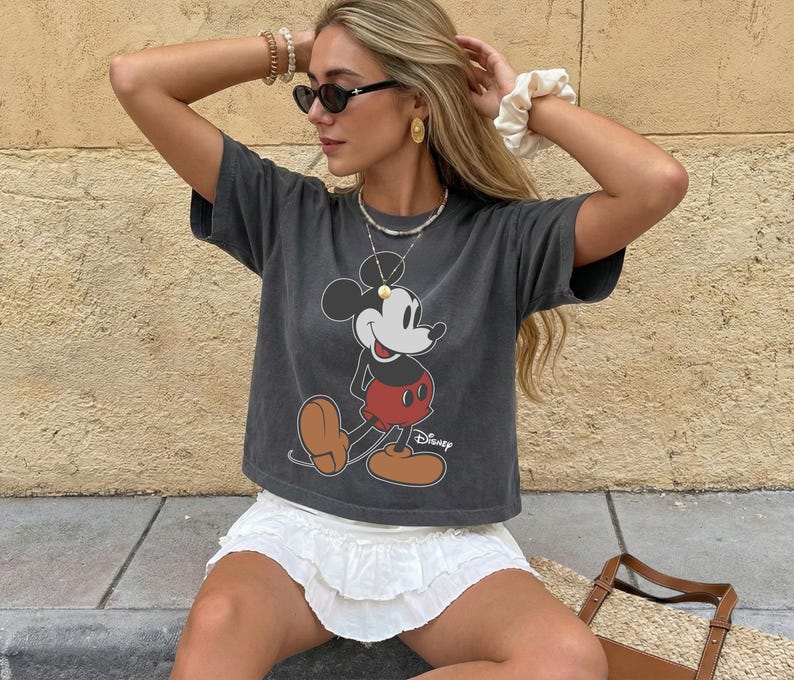 May include: A charcoal gray t-shirt featuring a classic Mickey Mouse graphic in red, black, and tan. The shirt is paired with a white ruffled skirt. The Disney logo is visible on the shirt.