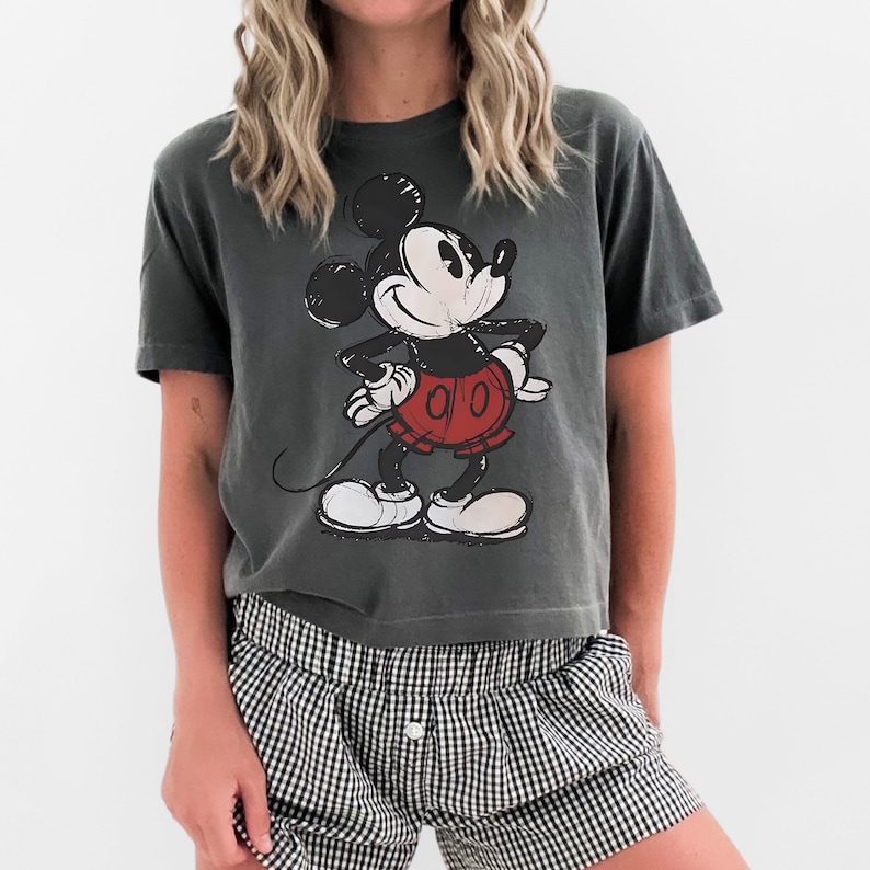 May include: Dark gray t-shirt featuring a distressed graphic of Mickey Mouse in his classic pose. Mickey is wearing red shorts, white gloves, and shoes. The shirt is paired with black and white checkered shorts.