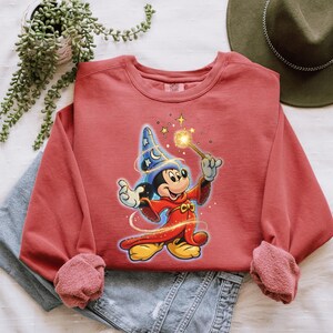 May include: A red-orange sweatshirt featuring a graphic of Mickey Mouse dressed as a wizard, holding a glowing wand. The design includes a blue wizard hat with stars and moons. The sweatshirt has long sleeves and a crew neck.