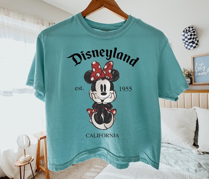 K&ouml;nnte beinhalten: Ein t&uuml;rkisfarbenes T-Shirt mit einem Minnie Mouse-Motiv mit roter Schleife und dem Text "Disneyland est. 1955 CALIFORNIA". Das T-Shirt h&auml;ngt an einem Holzb&uuml;gel.