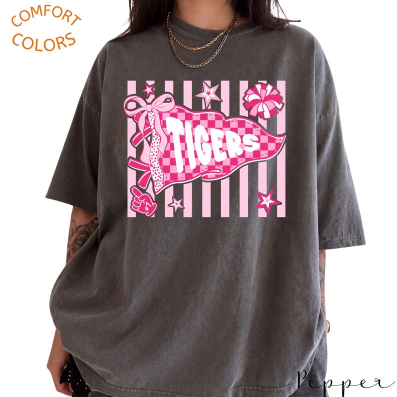 May include: A charcoal gray short-sleeve t-shirt featuring a graphic print of a pink checkered pennant with the text TIGERS, surrounded by pink stars, a pom-pom, and a foam finger, set against a background of vertical pink and white stripes.