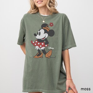 Może przedstawiać: Zielona koszulka w kolorze mchu z grafiką Myszki Minnie. Postać z kresk&oacute;wki ma na sobie czerwoną sukienkę w kropki, brązowe buty i kapelusz z kwiatkiem. Logo Disneya jest wydrukowane poniżej. Koszulka ma kr&oacute;tkie rękawy i luźny kr&oacute;j.