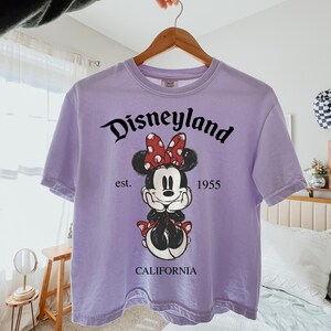 K&ouml;nnte beinhalten: Ein lila T-Shirt mit einem Minnie Mouse-Motiv. Das Design enth&auml;lt den Text "Disneyland" in schwarzer Schrift, "est. 1955" und "CALIFORNIA". Minnie Mouse ist mit einer roten Polka-Dot-Schleife und einem schwarzen Outfit dargestellt.