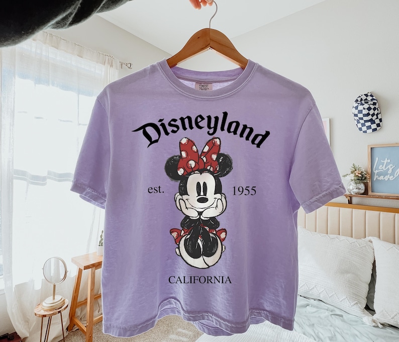K&ouml;nnte beinhalten: Ein lila T-Shirt mit einem Minnie Mouse-Motiv. Das Design enth&auml;lt den Text "Disneyland" in schwarzer Schrift, "est. 1955" und "CALIFORNIA". Minnie Mouse ist mit einer roten Polka-Dot-Schleife und einem schwarzen Outfit dargestellt.
