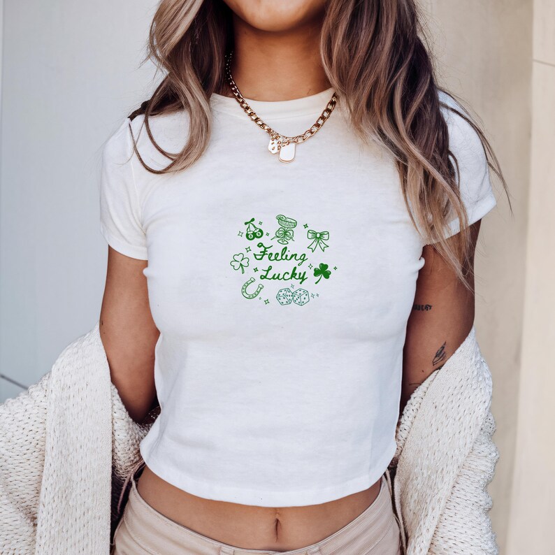 May include: White crop top with green "Feeling Lucky" graphic. The design includes a horseshoe, shamrocks, dice, and other St. Patrick's Day themed elements. The shirt is short-sleeved and has a crew neck.