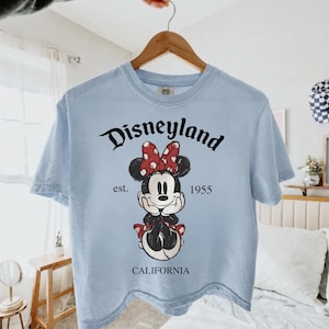 K&ouml;nnte beinhalten: Hellblaues T-Shirt mit einer Minnie Mouse-Grafik, einer roten Schleife und dem Text "Disneyland California 1955". Das T-Shirt h&auml;ngt an einem Holzb&uuml;gel.