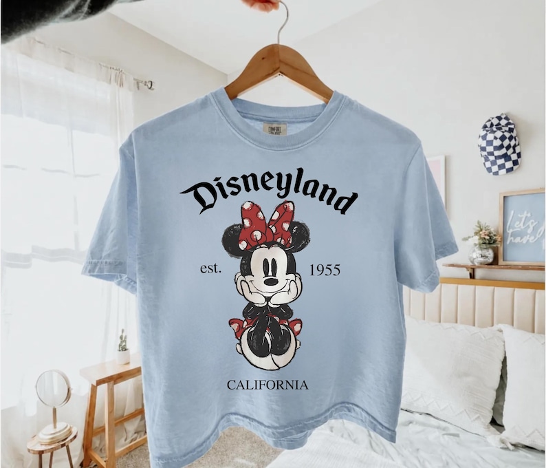 K&ouml;nnte beinhalten: Hellblaues T-Shirt mit einer Minnie Mouse-Grafik, einer roten Schleife und dem Text "Disneyland California 1955". Das T-Shirt h&auml;ngt an einem Holzb&uuml;gel.