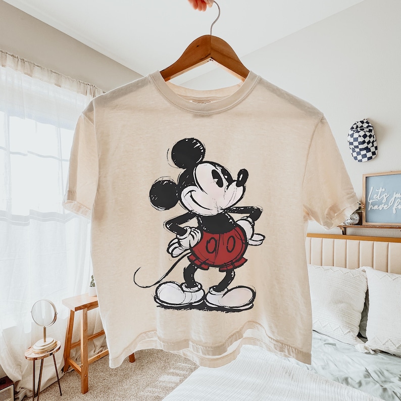 May include: A cream-colored t-shirt featuring a black and red graphic of Mickey Mouse. The cartoon character is in a classic pose with a smiling expression. The shirt is hanging on a wooden hanger.