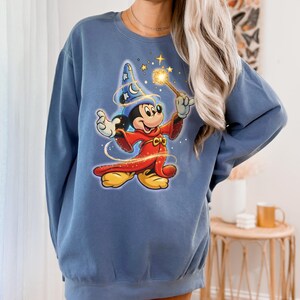 May include: A blue sweatshirt featuring a graphic of Mickey Mouse dressed as a sorcerer, holding a glowing wand. Mickey is wearing a blue wizard hat with stars and a crescent moon. The sweatshirt is a solid blue color.