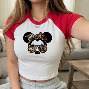 Op de afbeelding: Wit en rood raglan crop top met een Minnie Mouse afbeelding met een zonnebril en strik met luipaardprint. De top heeft korte mouwen en een ronde halslijn. Een casual stijl.