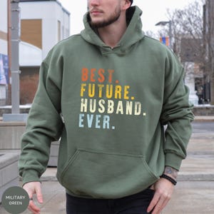 May include: Military green hoodie with the text "BEST. FUTURE. HUSBAND. EVER." in orange, yellow, white, and blue. The hoodie has a front pocket and a drawstring hood. The text is in a retro font.