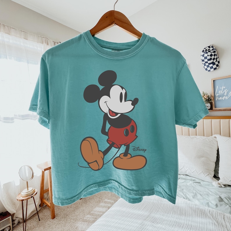 May include: Teal-colored t-shirt featuring a graphic of Mickey Mouse. The cartoon character is depicted in his classic outfit: red shorts, large yellow shoes, and white gloves. The Disney logo is printed below the character.