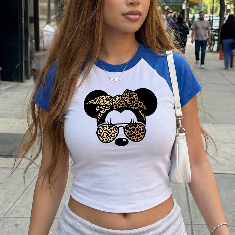 Op de afbeelding: Witte crop top met blauwe mouwen met een Minnie Mouse-afbeelding met een zonnebril en een luipaardprint strik. De shirt heeft een baseball-t-shirt-stijl.