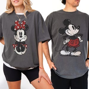 以下が含まれることがあります： ディズニーキャラクターが描かれたチャコールグレーのTシャツ2枚。1枚は赤いリボンをつけたミニーマウス、もう1枚は赤いパンツのミッキーマウス。どちらのシャツもヴィンテージ風のダメージ加工が施されています。