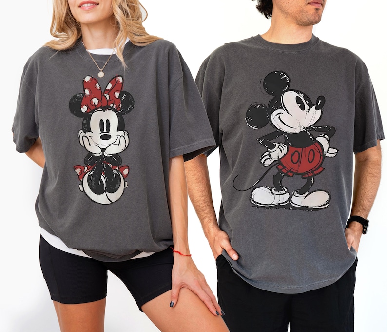 以下が含まれることがあります： ディズニーキャラクターが描かれたチャコールグレーのTシャツ2枚。1枚は赤いリボンをつけたミニーマウス、もう1枚は赤いパンツのミッキーマウス。どちらのシャツもヴィンテージ風のダメージ加工が施されています。