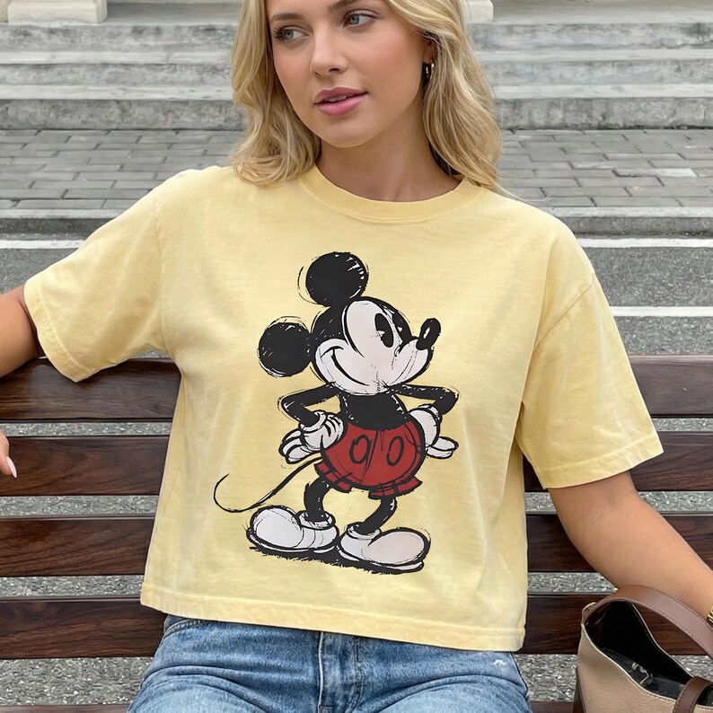 May include: A yellow t-shirt featuring a distressed graphic of Mickey Mouse in a classic pose. The cartoon character is depicted in black, white, and red. The shirt has short sleeves and a crew neck.