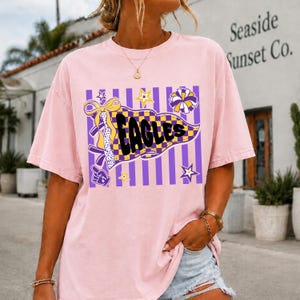 May include: A light pink short-sleeve t-shirt featuring a graphic print of a yellow and purple checkered pennant with the text "EAGLES" in black. The design includes purple vertical stripes, a yellow bow, stars, and a foam finger icon.