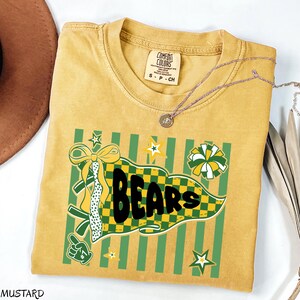 May include: A mustard yellow short-sleeve t-shirt with a Comfort Colors label. The graphic features a green and yellow checkered pennant with the word BEARS in black, a yellow bow, a green foam finger, stars, and a pom-pom on a striped background.